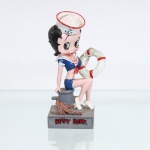 BRINQUEDO / COLECIONISMO. Estatueta decorativa colecionável em resina policromada figurando a personagem Betty Boop vestida como "Marinheira" / "Maruja". Coleção oficial licenciada, final do século XX ou início do século XXI. A icônica personagem surge representada sentada de perfil sobre um cabeço de amarração(bita) cinza de cais portuário, posicionado sobre base retangular em tom cinza-cimento. Apresenta-se com o rosto voltado para a frente, ostentando seus tradicionais grandes olhos verdes e brincos de argola dourados. Traja uniforme estilizado de marinheira composto por top curto decotado azul-marinho com debruns brancos, gola marinheiro (gola flutuante) caída nas costas, lenço frontal com amarração em laço vermelho e shorts curto combinando em azul-marinho. Sobre a cabeça, ostenta um clássico quepe de marinheiro(marujo) branco com debrum superior vermelho e estampa frontal de uma âncora azul. A figura apoia a perna esquerda flexionada e segura com o braço esquerdo uma boia salva-vidas circular nas cores branca e vermelha. A composição da base inclui uma corda/amarriga náutica de esparto marrom texturizada enrolada ao pé do cabeço. Calça sapatos de salto alto na cor branca.Na face frontal da base, inscrição em relevo com o nome da personagem "BETTY BOOP", destacado em esmalte vermelho com contorno preto. No reverso da base, apresenta inscrição de direitos autorais e licenciamento oficial gravada: "KFS/FS TM Hearst/FS". Medidas: Alt.: x Comp.: x Larg.: cm.