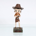 BRINQUEDO / COLECIONISMO. Estatueta decorativa colecionável em resina policromada figurando a personagem Betty Boop vestida como "Aventureira" / "Exploradora de Safari". Coleção oficial licenciada, final do século XX ou início do século XXI. A icônica personagem surge representada em pé sobre base retangular texturizada em tom verde-oliva escuro que simula um terreno de vegetação ou capim rasteiro. Apresenta-se em posição frontal, ostentando seus tradicionais grandes olhos verdes e brincos de argola na cor dourada. Traja indumentária de safari composta por camisa de mangas curtas cáqui/marrom-clara com bolsos frontais e amarração na cintura revelando o abdômen, saia curta de corte irregular/assimétrico em tom marrom-escuro com cinto preto, e botas de cano curto combinando. Sobre a cabeça, ostenta grande chapéu de explorador de abas largas texturizado, ornado por cordão trançado marrom em todo o contorno da copa. A figura segura com ambas as mãos um binóculo na cor preta à frente do peito, em posição de observação.Na face frontal da base, inscrição em relevo com o nome da personagem "BETTY BOOP", destacado em esmalte vermelho com contorno preto. No reverso da base, apresenta inscrição de direitos autorais e licenciamento oficial gravada: "KFS/FS TM Hearst/FS". Medidas: Alt.: x Comp.: x Larg.: cm.