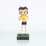 BRINQUEDO / COLECIONISMO. Estatueta decorativa colecionável em resina policromada figurando a personagem Betty Boop vestida como "Jogadora de Futebol". Coleção oficial licenciada, final do século XX ou início do século XXI.A icônica personagem surge representada em pé sobre base retangular texturizada em tom verde-escuro que simula um gramado esportivo. Apresenta-se em posição frontal, com a mão esquerda apoiada na cintura e ostentando seus tradicionais grandes olhos verdes. Traja uniforme clássico de futebol com as cores da seleção brasileira, composto por camisa amarela de gola em "V" com debruns verdes nas mangas e o número "11" estampado em verde no peito e nas costas, calção curto azul-marinho com listras brancas laterais e numeração correspondente, meias longas(meiões) brancas texturizadas e chuteiras na cor azul-marinho. Sob o braço direito, a figura carrega uma bola de futebol estilizada em tons de branco e amarelo.Na face frontal da base, inscrição em relevo com o nome da personagem "BETTY BOOP", destacado em esmalte vermelho com contorno preto. No reverso da base, apresenta inscrição de direitos autorais e licenciamento oficial gravada: "KFS/FS TM Hearst/FS". Medidas: Alt.: x Comp.: x Larg.: cm.