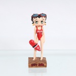Estatueta decorativa / boneca colecionável em resina policromada figurando a personagem Betty Boop vestida como "Salva-Vidas" / "Guarda-Vidas"(Lifeguard). Coleção oficial licenciada, final do século XX ou início do século XXI. A icônica personagem surge representada descalça e em pé sobre base quadrada texturizada em tom marrom-escuro que simula uma superfície de areia molhada de praia. Apresenta-se em posição frontal, ostentando seus tradicionais grandes olhos verdes e brincos de argola na cor amarela. Traja maiô clássico de peça única na cor vermelha, trazendo estampado no peito a inscrição em inglês "LIFEGUARD" em letras brancas, disposta acima de um pequeno emblema circular com uma cruz médica branca. Sobre a cabeça, traz óculos de sol estilizados com armação vermelha e lentes azuis, posicionados acima da testa. Com a mão direita estendida para baixo, segura um flutuador de salvamento tipo life-ring ou rescue tube na cor vermelha, ornamentado por uma faixa central branca contendo o mesmo emblema de cruz médica.Na face frontal da base, inscrição em relevo com o nome da personagem "BETTY BOOP", destacado em esmalte vermelho com contorno preto. No reverso da base, apresenta inscrição de direitos autorais e licenciamento oficial gravada: "KFS/FS TM Hearst/FS".