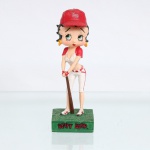 BRINQUEDO / COLECIONISMO. Estatueta decorativa / boneca colecionável em resina policromada figurando a personagem Betty Boop vestida como "Jogadora de Beisebol". Coleção oficial licenciada, final do século XX ou início do século XXI. A icônica personagem surge representada em pé sobre base retangular texturizada em tom verde-escuro que simula um gramado esportivo. Apresenta-se em posição frontal, com o corpo ligeiramente inclinado à frente em postura de prontidão. Ostenta seus tradicionais grandes olhos verdes e brincos de argola amarelos. Traja uniforme estilizado de beisebol composto por camisa curta estilo cropped na cor branca com mangas curtas vermelhas, calça curta modelo knickers(abaixo do joelho) branca com frisos laterais vermelhos, cinto vermelho e luvas de rebatida combinando. Sobre a cabeça, ostenta um clássico boné de beisebol vermelho voltado para a frente, decorado na porção frontal pelo monograma bordado estilizado "BB" em relevo branco. Com ambas as mãos enluvadas, a figura apoia verticalmente à frente do corpo um bastão de beisebol de madeira texturizado em tom marrom-escuro. Calça tênis esportivos brancos com travas e detalhes em vermelho.Na face frontal da base, inscrição em relevo com o nome da personagem "BETTY BOOP", destacado em esmalte vermelho com contorno preto. No reverso da base, apresenta inscrição de direitos autorais e licenciamento oficial gravada: "KFS/FS TM Hearst/FS". Medidas: Alt.: x Comp.: x Larg.: cm.