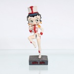 BRINQUEDO / COLECIONISMO. Estatueta decorativa colecionável em resina policromada figurando a personagem Betty Boop vestida como "Majorete" / "Baliza de Fanfarra". Coleção oficial licenciada, final do século XX ou início do século XXI. A icônica personagem surge representada em posição dinâmica de marcha, com a perna direita flexionada e suspensa no ar, sobre base quadrada texturizada em tom cinza-escuro. Apresenta-se em posição frontal, com os tradicionais grandes olhos verdes e brincos de argola na cor amarela. Traja uniforme militar estilizado de parada composto por jaqueta branca acinturada com alamares franceses horizontais, botões e debruns na cor vermelha, dragonas com franjas douradas nos ombros, punhos vermelhos e shorts curto branco combinando. Sobre a cabeça, ostenta um clássico chapéu alto de desfile(shako) na cor branca, guarnecido de aba azul, cordão vermelho na base e um imponente pompom ou pluma(plumache) vertical na cor vermelha. Com a mão direita flexionada junto ao peito, a figura segura um bastão de comando prateado, mantendo a mão esquerda apoiada firmemente na cintura. Calça botas de cano alto brancas com amarrações frontais e franjas decorativas pendentes na cor vermelha.Na face frontal da base, inscrição em relevo com o nome da personagem "BETTY BOOP", destacado em esmalte vermelho com contorno preto. No reverso da base, apresenta inscrição de direitos autorais e licenciamento oficial gravada: "KFS/FS TM Hearst/FS". Medidas: Alt.: x Comp.: x Larg.: cm.