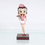 BRINQUEDO / COLECIONISMO. Estatueta decorativa colecionável em resina policromada figurando a personagem Betty Boop vestida como 'Garçonete de Lanchonete Retrô"(Diner Waitress). Coleção oficial licenciada, final do século XX ou início do século XXI. A icônica personagem surge representada em pé com as pernas cruzadas sobre base retangular cinza-escura de textura áspera. Apresenta-se em posição frontal, com a mão esquerda apoiada na cintura e ostentando seus tradicionais grandes olhos verdes e brincos de argola na cor vermelha. Traja uniforme retrô de atendente de lanchonete dos anos 1950 na cor rosa, composto por vestido curto acinturado com lapelas e debruns pretos nas mangas, avental curto branco com amarrações em laço no reverso e detalhes em vermelho na cintura. Sobre a cabeça, ostenta um clássico casquete rosa com debrum vermelho, decorado na porção frontal pelo nome estilizado "Betty". Com a mão direita estendida à frente, a figura equilibra uma bandeja cinza redonda contendo uma xícara de café dourada e um pequeno recipiente prateado. Calça sapatos clássicos de salto alto na cor vermelha.Na face frontal da base, inscrição em relevo com o nome da personagem "BETTY BOOP", destacado em esmalte vermelho com contorno preto. No reverso da base, apresenta inscrição de direitos autorais e licenciamento oficial gravada: "KFS/FS TM Hearst/FS". Medidas: Alt.: x Comp.: x Larg.: cm.