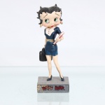 BRINQUEDO / COLECIONISMO. Estatueta decorativa colecionável em resina policromada figurando a personagem Betty Boop vestida como "Executiv"a / "Mulher de Negócios (Businesswoman). Coleção oficial licenciada, final do século XX ou início do século XXI. A icônica personagem surge representada em pé sobre base quadrada cinza-escura de textura áspera. Apresenta-se em posição frontal, ostentando seus tradicionais grandes olhos verdes e brincos de argola na cor dourada. Traja indumentária social formal e justa na cor azul-marinho, composta por vestido curto estilo tailleur ou saia e blusa texturizada com decote acentuado transpassado por gola e lapelas brancas com detalhe de abotoamento triplo em preto. O traje é acinturado por um cinto dourado espesso. Com a mão esquerda flexionada junto ao peito, carrega uma pasta de relatórios ou livro na cor vermelha; com o braço direito estendido para baixo, segura a alça de uma maleta executiva / pasta de documentos na cor preta. Calça sapatos clássicos de salto alto no modelo scarpin também em azul-marinho.Na face frontal da base, inscrição em relevo com o nome da personagem "BETTY BOOP", destacado em esmalte vermelho com contorno preto. No reverso da base, apresenta inscrição de direitos autorais e licenciamento oficial gravada:"KFS/FS TM Hearst/FS". Medidas: Alt.: x Comp.: x Larg.: cm.