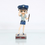 BRINQUEDO / COLECIONISMO. Estatueta decorativa colecionável em resina policromada figurando a personagem Betty Boop vestida como "Policial de Trânsito". Coleção oficial licenciada, final do século XX ou início do século XXI. A icônica personagem surge representada em pé sobre base retangular cinza que simula uma faixa de pedestres zebrada com listras brancas. Apresenta-se com o corpo projetado para trás e a mão direita elevada à altura da testa em um gesto de observação / proteção contra a luz solar, ostentando seus tradicionais grandes olhos verdes e brincos de argola amarelos. Traja uniforme estilizado composto por camisa de mangas curtas em tom azul-esverdeado claro com decote acentuado e lapelas abertas, minissaia justa em azul-escaro/marinho com cinto preto de fivela prateada e coldre lateral. Sobre a cabeça, ostenta um quepe policial azul-escuro com aba preta e insígnia prateada de escudo centralizada na copa. Com a mão esquerda estendida lateralmente, segura um cassetete/bastão policial na cor preta em posição vertical. Calça sapatos clássicos de salto alto na cor azul.Na face frontal da base, inscrição em relevo com o nome da personagem "BETTY BOOP", destacado em esmalte vermelho com contorno preto. No reverso da base, apresenta inscrição de direitos autorais e licenciamento oficial gravada: "KFS/FS TM Hearst/FS". Medidas: Alt.: x Comp.: x Larg.: cm.