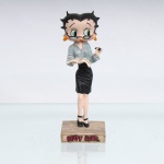BRINQUEDO / COLECIONISMO. Estatueta decorativa colecionável em resina policromada figurando a personagem Betty Boop vestida como "Professora". Coleção oficial licenciada, final do século XX ou início do século XXI. A icônica personagem surge representada em pé sobre base retangular ocre-clara com textura incisa que simula tábuas de um piso de madeira. Apresenta-se em posição frontal, ostentando seus tradicionais grandes olhos verdes e brincos de argola na cor amarela, trazendo como diferencial um par de óculos de leitura de armação preta posicionado sobre o nariz. Traja indumentária social clássica composta por camisa de mangas compridas azul-claro com abotoamento frontal e gola aberta, combinada com uma saia secretária justa de cintura alta na cor preta. Com a mão esquerda flexionada, a figura segura uma caneta preta e prateada; com a mão direita estendida à frente, apoia um livro ou caderno aberto em posição de leitura. Calça sapatos clássicos de salto alto com tira no peito do pé(mary jane) na cor preta.Na face frontal da base, inscrição em relevo com o nome da personagem "BETTY BOOP", destacado em esmalte vermelho com contorno preto. No reverso da base, apresenta inscrição de direitos autorais e licenciamento oficial gravada: "KFS/FS TM Hearst/FS". Medidas: Alt.: x Comp.: x Larg.: cm.