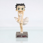 BRINQUEDO / COLECIONISMO. Estatueta decorativa colecionável em resina policromada figurando a personagem Betty Boop em sua clássica pose inspirada em Marilyn Monroe, com o vestido esvoaçante. Coleção oficial licenciada, final do século XX ou início do século XXI. A icônica personagem surge representada em pé sobre base quadrada cinza-escura com textura de grelha em relevo que simula um bueiro de ventilação urbana. Apresenta-se com o corpo levemente inclinado à frente e as mãos unidas sobre as coxas em atitude de modéstia, contendo o panejamento da saia. Ostenta seus tradicionais grandes olhos verdes, brincos de argola amarelos e uma liga vermelha com coração na coxa direita. Traja clássico vestido de frente única tomara-que-caia na cor branca, cuja saia plissada surge amplamente esvoaçante e armada nas laterais por uma simulação de corrente de ar. Calça sapatos de salto alto também na cor branca.Na face frontal da base, inscrição em relevo com o nome da personagem "BETTY BOOP", destacado em esmalte vermelho com contorno preto. No reverso da base, apresenta inscrição de direitos autorais e licenciamento oficial gravada: "KFS/FS TM Hearst/FS". Medidas: Alt.: x Comp.: x Larg.: cm.