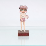 BRINQUEDO / COLECIONISMO. Estatueta decorativa colecionável em resina policromada figurando a personagem Betty Boop vestida como "Jogadora de Tênis". Coleção oficial licenciada, final do século XX ou início do século XXI. A icônica personagem surge representada em pé sobre base quadrada marrom-terrosa que simula um piso de quadra de saibro. Apresenta-se em posição frontal, com os tradicionais grandes olhos verdes e brincos de argola na cor rosa. Traja uniforme esportivo composto por camisa polo branca de mangas curtas e gola aberta, combinada com minissaia pregueada na cor rosa. Sobre a cabeça, ostenta uma viseira de tênis rosa texturizada, decorada na porção frontal pelo nome estilizado "Betty". Com a mão esquerda, a figura segura uma raquete de tênis rosa com detalhes brancos na rede; com a mão direita flexionada junto à cintura, segura uma bola de tênis na cor amarela/laranja. Calça meias curtas e tênis esportivos na cor branca.Na face frontal da base, inscrição em relevo com o nome da personagem "BETTY BOOP", destacado em esmalte vermelho com contorno preto. No reverso da base, apresenta inscrição de direitos autorais e licenciamento oficial gravada: "KFS/FS TM Hearst/FS". Medidas: Alt.: x Comp.: x Larg.: cm.