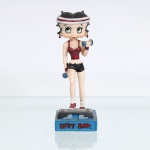 BRINQUEDO / COLECIONISMO. Estatueta decorativa colecionável em resina policromada figurando a personagem Betty Boop Praticando Fitness / Aeróbica. Distribuição oficial licenciada pela Editora Salvat do Brasil Ltda. (Edição nº 22). A icônica personagem surge representada em ação, executando exercícios físicos sobre uma plataforma de step cinza e preta. Apresenta-se com seus característicos grandes olhos verdes e brincos de argola na cor vermelha, trazendo a cabeça cingida por uma faixa de suor(headband) nas cores vermelha e branca. Traja conjunto esportivo composto por top de alças em tom vermelho-grená metálico e shorts curto(hot pants) na cor preta. Segura um par de halteres(pesos) azuis, um em cada mão, elevando o braço esquerdo em flexão. Calça meias brancas de cano médio e tênis esportivos vermelhos e brancos.  Fixada sobre uma base retangular texturizada azul-turquesa. Na face frontal da base, inscrição em relevo com o nome da personagem "BETTY BOOP", destacado em esmalte vermelho com contorno preto. No reverso da base, apresenta inscrição de direitos autorais e licenciamento oficial gravada: "KFS/FS TM Hearst/FS". Sob o fundo da base, apresenta etiqueta comercial completa de importação com dados da distribuidora nacional, CNPJ e a indicação técnica da coleção: "For edition 22 - Artigo para colecionistas. Não é um brinquedo". Medidas: Alt.: x Comp.: x Larg.: