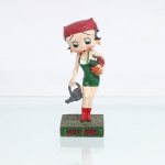 BRINQUEDO / COLECIONISMO. Estatueta decorativa colecionável em resina policromada figurando a personagem Betty Boop vestida como Jardineira / Camponesa. Coleção oficial licenciada, final do século XX ou início do século XXI. A icônica personagem surge representada em pé sobre base retangular texturizada em tom verde-musgo simulando gramado. Apresenta-se com o corpo levemente inclinado à frente, ostentando seus tradicionais grandes olhos verdes e brincos de argola amarelos. Traja jardineira jeans curta na cor verde, sobreposta a uma blusa regata branca, complementada por lenço amarrado na cabeça(bandana) na cor vermelha com nó em seu reverso. Com a mão direita voltada para baixo, segura um regador de plantas metálico estilizado na cor cinza-escura; sob o braço esquerdo, carrega uma cesta de vime preenchida por hortaliças e flores policromadas em tons de vermelho, verde e laranja. Calça galochas de cano médio na cor vermelha. Na face frontal da base, inscrição em relevo com o nome da personagem "BETTY BOOP", destacado em esmalte vermelho com contorno preto. No reverso da base, apresenta inscrição de direitos autorais e licenciamento oficial gravada: "KFS/FS TM Hearst/FS". Medidas: Alt.: x Comp.: x Larg.: cm.