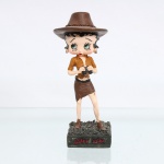BRINQUEDO / COLECIONISMO. Estatueta decorativa colecionável em resina policromada figurando a personagem Betty Boop vestida como "Exploradora de Safari" / "Aventureira". Coleção oficial licenciada, final do século XX ou início do século XXI. A icônica personagem surge representada em pé sobre base retangular texturizada em tom verde-oliva escuro que simula um terreno de vegetação ou capim rasteiro. Apresenta-se em posição frontal, ostentando seus tradicionais grandes olhos verdes e brincos de argola na cor dourada. Traja indumentária de safari composta por camisa de mangas curtas ocre/cáqui com bolsos frontais e lapelas, aberta na região do colo, combinada com uma minissaia justa de corte irregular/assimétrico em tom marrom-escuro com cinto preto. Sobre a cabeça, ostenta um grande chapéu de explorador de abas largas texturizado na cor marrom, ornado por um cordão trançado em todo o contorno da copa com amarração posterior em laço. A figura segura com ambas as mãos um binóculo na cor preta à frente do peito, em posição de observação. Calça botas de amarrar com cano curto em tom marrom.Na face frontal da base, inscrição em relevo com o nome da personagem "BETTY BOOP", destacado em esmalte vermelho com contorno preto. No reverso da base, apresenta inscrição de direitos autorais e licenciamento oficial gravada: "KFS/FS TM Hearst/FS". Medidas: Alt.: x Comp.: x Larg.: cm.
