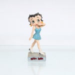 BRINQUEDO / COLECIONISMO. Estatueta decorativa colecionável em resina policromada figurando a personagem Betty Boop como "Patinadora Artística no Gelo". Coleção oficial licenciada, final do século XX ou início do século XXI. A icônica personagem surge representada em posição dinâmica de deslizamento sobre base retangular cinza-clara que simula uma pista de gelo texturizada. Apresenta-se com o corpo levemente inclinado à frente, com o braço esquerdo flexionado levando a mão delicadamente próxima à boca em gesto expressivo, ostentando seus tradicionais grandes olhos verdes e brincos de argola na cor amarela. Traja vestido curto de patinação de alças finas na cor azul-turquesa, apresentando corpete ajustado e saia curta rodada com panejamento plissado em movimento. Calça botas de patinação de cano médio na cor branca com amarrações incisas, equipadas com lâminas metálicas prateadas e suportes pretos fixados ao solado.Na face frontal da base, inscrição em relevo com o nome da personagem "BETTY BOOP", destacado em esmalte vermelho com contorno preto. No reverso da base, apresenta inscrição de direitos autorais e licenciamento oficial carimbada em preto: "KFS/FS TM Hearst/FS". Medidas: Alt.: x Comp.: x Larg.: cm.