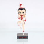 BRINQUEDO / COLECIONISMO. Estatueta decorativa colecionável em resina policromada figurando a personagem Betty Boop vestida como "Majorete" / "Baliza de Fanfarra". Coleção oficial licenciada, final do século XX ou início do século XXI. A icônica personagem surge representada em posição dinâmica de marcha, com a perna direita flexionada e suspensa no ar, sobre base quadrada texturizada em tom cinza-escuro. Apresenta-se em posição frontal, ostentando seus tradicionais grandes olhos verdes e brincos de argola na cor amarela. Traja uniforme militar estilizado de desfile composto por jaqueta branca curta(cropped) com alamares franceses horizontais, botões e punhos na cor vermelha, dragonas com franjas douradas nos ombros e shorts curto branco combinando com detalhe em vermelho na lateral. Sobre a cabeça, ostenta um clássico chapéu alto de desfile(shako) na cor branca, guarnecido de aba azul-escura, cordão vermelho na base e um imponente pompom ou pluma(plumache) vertical na cor vermelha. Com a mão direita flexionada junto ao peito, a figura segura um bastão de comando prateado, mantendo a mão esquerda apoiada firmemente na cintura. Calça botas de cano alto brancas com amarrações frontais e franjas decorativas pendentes na cor vermelha.Na face frontal da base, inscrição em relevo com o nome da personagem "BETTY BOOP", destacado em esmalte vermelho com contorno preto. No reverso da base, apresenta inscrição de direitos autorais e licenciamento oficial gravada: "KFS/FS TM Hearst/FS". Medidas: Alt.: x Comp.: x Larg.: cm.