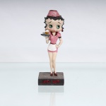 BRINQUEDO / COLECIONISMO. Estatueta decorativa / boneca colecionável em resina policromada figurando a personagem Betty Boop vestida como "Garçonete de Lanchonete Retrô"(Diner Waitress). Coleção oficial licenciada, final do século XX ou início do século XXI. A icônica personagem surge representada em pé sobre base quadrada marrom-escura de textura áspera. Apresenta-se em posição frontal, com a mão esquerda apoiada na cintura e ostentando seus tradicionais grandes olhos verdes e brincos de argola na cor vermelha. Traja uniforme retrô de atendente de lanchonete na cor rosa, composto por vestido curto acinturado com lapelas e debruns marons/pretos nas mangas, avental curto branco com amarrações em laço no reverso e detalhes em vermelho na cintura. Sobre a cabeça, ostenta um clássico casquete(pillbox hat) rosa com debrum marrom, decorado na porção frontal pelo nome estilizado "Betty". Com a mão direita estendida à frente, a figura equilibra uma bandeja cinza redonda contendo uma xícara de café dourada e um pequeno recipiente prateado. Calça sapatos clássicos de salto alto na cor vermelha.Na face frontal da base, inscrição em relevo com o nome da personagem "BETTY BOOP", destacado em esmalte vermelho com contorno preto. No reverso da base, apresenta inscrição de direitos autorais e licenciamento oficial gravada: "KFS/FS TM Hearst/FS". Medidas: Alt.: x Comp.: x Larg.: cm.