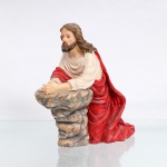 Figura devocional executada em resina policromada, representando Jesus Cristo em oração no Getsêmani. Escultura moldada representando Cristo ajoelhado em oração, com as mãos unidas e apoiadas sobre formação rochosa, iconografia alusiva à "Agonia de Jesus no Jardim do Getsêmani", episódio que antecede sua prisão e a Paixão. Apresenta cabelos longos e barba, trajando túnica em tonalidade marfim e manto vermelho disposto em pregas, com base integrada simulando rocha. Sem marca de manufatura. Medidas: Alt.: 18,5 x Comp.: 18,5 x Larg.: 10,5cm.
