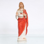 Imagem devocional do Sagrado Coração de Jesus em porcelana policromada. Figura moldada representando Jesus Cristo sob a invocação do Sagrado Coração, de pé sobre base oitavada. Apresenta cabelos longos e barba, túnica branca cingida à cintura e manto vermelho disposto sobre o ombro esquerdo. Ao centro do peito figura o Sagrado Coração, encimado por cruz e envolvido pela coroa de espinhos; a mão direita encontra-se elevada em gesto de bênção, exibindo a chaga da Crucificação, igualmente assinalada na mão esquerda. Policromia em vermelho, verde, castanho e dourado. Base vazada, sem marca de manufatura. Medidas: Alt.: 21 x Comp.: 6 x Larg.: 5cm.