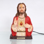 Busto devocional do Sagrado Coração de Jesus, eletrificado, executado em resina policromada. Imagem moldada representando Jesus Cristo sob a invocação do Sagrado Coração, em meio-corpo, com a mão direita elevada em gesto de bênção e a esquerda indicando o coração disposto sobre o peito. O atributo central apresenta coração flamejante, encimado por cruz e envolvido pela coroa de espinhos, sobre resplendor translúcido de disposição radial. Cristo veste túnica em tonalidade marfim, cingida à cintura, e manto vermelho. Assenta sobre base retangular em tom castanho, provida de placa metálica com a inscrição Sagrado Coração de Jesus e sistema elétrico destinado à iluminação interna. Sem marca de manufatura. Medidas: Alt.: 21,5 x Comp.: 14 x Larg.: 8,5cm.