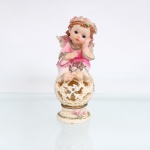 Figura decorativa de anjo infantil com mecanismo luminoso, em resina policromada; moldada representando anjo de feições infantis, sentado sobre esfera vazada, com asas, cabelos ondulados e coroa de flores. Traja veste em tonalidades rosa e marfim, ornamentada por motivos florais em relevo, folhagens, aplicações de brilho e realces dourados. A esfera apresenta composição vazada de volutas, folhas de acanto, flores e reservas cruciformes, assentando sobre pedestal circular moldurado, igualmente guarnecido por rosas e elementos fitomórficos. Base provida de compartimento para pilhas, interruptor e mecanismo interno destinado à iluminação através dos vazados. Sem marca de manufatura. Medidas: Alt.: 19 x Comp.: 7 x Larg.: 7,5cm.