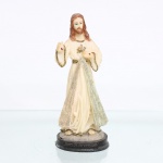 Imagem devocional de Jesus Misericordioso em resina policromada. Figura moldada representando Jesus Cristo segundo a iconografia da Divina Misericórdia, de pé, com a mão direita elevada em gesto de bênção e a esquerda direcionada ao peito, trazendo nas palmas as chagas da Crucificação. Traja túnica longa em tonalidade marfim, cingida à cintura, com aplicações douradas e prateadas. Assenta sobre base oval em tom escurecido, com inscrição moldada "Jesus Misericordioso"; junto aos pés, lê-se parcialmente a fórmula devocional "Jesus, confio em Vós", associada à imagem da Divina Misericórdia. Essa representação tem origem nas visões relatadas por Santa Faustina Kowalska em 1931, nas quais Cristo teria solicitado a execução de uma imagem acompanhada da inscrição "Jesus, eu confio em Vós". A iconografia difundiu-se internacionalmente no culto católico ao longo do século XX. Medidas: Alt.: 5 x Comp.: 6 x Larg.: 5,5cm.