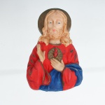 Busto devocional do Sagrado Coração de Jesus em resina policromada. Imagem moldada representando Jesus Cristo em meio-corpo, com nimbo circular, mão direita elevada em gesto de bênção e esquerda disposta junto ao peito. Ao centro figura o Sagrado Coração, flamejante e envolvido pela coroa de espinhos, inserido em resplendor circular com raios moldados. Traja túnica vermelha e manto azul, com contornos realçados em dourado. Verso plano, sem marca de manufatura ou autoria visível. A composição segue a iconografia católica do Sagrado Coração, difundida na imaginária devocional doméstica dos séculos XIX e XX. Medidas: Alt.: 12 x Comp.: 8 x Larg.: 4cm.