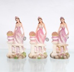 Três(03) figuras decorativas femininas em resina policromada, comemorativas de 15 anos. Figuras moldadas de composição idêntica, representando jovem em traje longo de tonalidades rosa e marfim, com ornamentação floral em relevo, aplicação de partículas brilhantes, tiara, brincos e colar com elemento facetado. Cada personagem apoia-se junto a balaustrada com vaso de flores, sobre base de aspecto rochoso e vegetação moldada. Apresentam mecanismo musical eletrônico instalado na base, acionado por interruptor e alimentado por pilhas. Cada exemplar conserva medalhão metálico suspenso com o numeral 15, referência aos quinze anos, indicando utilização como peça comemorativa ou lembrança de debutante. Medidas: Alt.: 17 x Comp.: 8,5 x Larg.: 7cm.