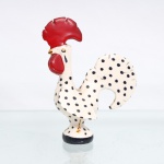 Escultura decorativa em faiança vidrada figurando o tradicional Galo de Barcelos, representada de perfil em sua modelagem clássica estilizada, assentada sobre base circular pintada em azul-cobalto escuro. O corpo apresenta fundo esmaltado na cor branca, inteiramente decorado por uma pintura manual de padrão de poás(pintas) em azul-escuro. Destacam-se em forte relevo a imponente crista e a barbela esmaltadas em vermelho-vivo, além dos olhos circulares detalhados em azul e amarelo, bico pintado em tom ocre; verso do corpo exibe as linhas sinuosas e vazadas da cauda arqueada características do icônico símbolo português. Medidas: Alt.: 14 x Comp.: 10 x Larg.: 6,5cm.