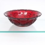 Fruteira/centro de mesa circular em vidro prensado na tonalidade vermelho-rubi profundo(Ruby Glass). Padrão geométrico moldado estilo Cape Cod, c. 1950/60. Formato hemisférico, de corpo robusto com bojo ricamente ornamentado em relevo no reverso por faixas de ogivas entrelaçadas, medalhões ovoides texturizados imitando bago de trigo e pequenas pontas de diamante e estrelas de quatro pontas. A borda superior exibe perfil ondulado lobado com acabamento liso e aba ligeiramente evertida, fundo externo assenta sobre base anelar, ostentando complexo medalhão central radiado com raios em ponta de lança e arabescos florais estilizados geometricamente.Medidas: Alt.: 8 x Diam.: 27 cm.