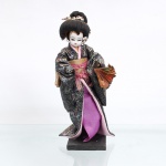 Boneca de coleção japonesa tradicional(Ningyo), figurando uma Geisha / dançarina clássica. Japão, segunda metade do século XX(c. 1960-1980). Representada em pé sobre base quadrangular de madeira pintada de preto. Apresenta cabeça moldada em gesso ou composição(gofun), com feições pintadas à mão detalhando olhos amendoados e lábios carmim, emoldurada por penteado tradicional elaborado em fios sintéticos pretos, ricamente ornamentado por presilhas e flores artificiais. Traja indumentária típica sobreposta composta por quimono de caimento fluido em tecido brocado escuro com fios metalizados furtacor em padronagem floral floral, subquimono em tom rosa, saia inferior em seda violeta, além de faixa abdominal(obi) dourada arrematada por laço e fitas cor-de-rosa em seu reverso. Em sua mão esquerda flexionada, segura um leque dobrável decorativo. Medidas: Alt.: x Comp.: x Larg.: cm.