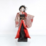 Boneca japonesa tradicional(Ningyo), figurando uma Geisha / dançarina clássica. Japão, segunda metade do século XX(c.1960-1980). Representada em pé sobre base quadrada de madeira pintada de preto. Apresenta cabeça moldada em gesso ou composição(gofun), com delicadas feições pintadas à mão ressaltando os olhos com delineado marcante e lábios carmim. O penteado tradicional é elaborado em fios sintéticos pretos amarrados em rabo de cavalo longo na parte posterior, ricamente ornamentado por presilhas metálicas e arranjos de flores artificiais brancas e vermelhas. Traja indumentária típica sobreposta composta por quimono de mangas largas em tecido brocado listrado nas cores prata e vermelho-coral com fios metalizados, subquimono com debrum vermelho, faixa abdominal(obi) preta arrematada por cordão dourado trançado em laço frontal e uma grande almofada protuberante escura em seu reverso. A saia interna longa estende-se até a base simulando o panejamento do traje tradicional em movimento. Medidas: Alt.: x Comp.: x Larg.: cm.