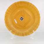Prato raso em vidro prensado de tonalidade âmbar, fundo texturizado, centro marcado por molduras concêntricas de contorno quadrilobado e aba delimitada por filetes circulares. Bordo ondulado. Conserva etiqueta original com as inscrições "Golden Pine by Wheaton  Made in Brazil", identificando a linha comercial e a manufatura brasileira Wheaton. Medida: Diam.: 29 cm.