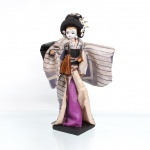 Boneca japonesa tradicional(Ningyo), figurando uma Geisha / dançarina clássica. Segunda metade do século XX(c. 1960-1980). A figura feminina surge representada em pé sobre base quadrada de madeira pintada de preto. Apresenta cabeça moldada em gesso ou composição gofun), com delicadas feições pintadas à mão ressaltando os olhos amendoados e lábios carmim. O penteado tradicional é elaborado em fios sintéticos pretos amarrados em coque alto, ornamentado por presilhas e flores artificiais brancas. Traja indumentária típica sobreposta composta por quimono de mangas largas em tecido brocado listrado nas cores prata e azul-violeta com fios metalizados e discretos grafismos geométricos em losango, subquimono com debrum em tom bege, faixa abdominal(obi) preta arrematada por laço de fita em seu reverso e saia inferior em seda violeta. Com a mão direita flexionada, a figura apoia verticalmente um tradicional guarda-sol / sombrinha de papel fechado em tom marrom. Medidas: Alt.: x Comp.: x Larg.: cm.