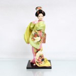 Boneca decorativa de estilo oriental / japonês(Ningyo), figurando uma Geisha tradicional. Manufatura moderna, final do século XX ou início do século XXI. Produzida na China para o mercado de exportação. Representada sobre base retangular de madeira escurecida, em uma pose de dança clássica com o corpo levemente sinuoso e as mãos elevadas à frente do tórax. Apresenta cabeça moldada em gesso, resina ou composição, com feições finamente pintadas à mão ressaltando os traços dos olhos e os lábios carmim. O penteado é elaborado em fios sintéticos pretos amarrados em coque alto, ornamentado por fitas e laços em tecidos azul e laranja com estampa floral. Traja indumentária típica sobreposta composta por quimono de mangas largas em tecido acetinado na cor verde-limão/pistache, ricamente decorado com estampas de peônias policromadas e uma garça branca de asas estendidas. O traje é complementado por subquimono rosa, saia inferior amarela e uma larga faixa abdominal(obi) rígida em tom coral com padronagem geométrica quadriculada de losangos dourados em seu reverso. Sob o fundo da base de madeira, apresenta etiqueta adesiva comercial impressa MADE IN CHINA. Medidas: Alt.: x Comp.: x Larg.: cm.