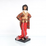 Boneca japonesa tradicional(Ningyo), representando uma jovem ou figura folclórica. Representada em pé sobre base quadrada de madeira escurecida, apresenta cabeça moldada em gesso ou composição(gofun), com feições pintadas à mão detalhando olhos amendoados e pequenos lábios vermelhos, cabelo curto e frisado confeccionado em mateial sintético, trazendo uma pequena fita rosa na lateral esquerda da franja lisa. Traja indumentária típica sobreposta composta por quimono de mangas curtas e caimento estruturado em tecido brocado encorpado na cor terracota/laranja-queimado com padronagem geométrica e fios metalizados, subquimono com gola transpassada branca e bege, e faixa abdominal(obi) em fibra texturizada bege-claro, arrematada por laço plano em seu reverso; saia inferior longa estende-se em panejamento fluido de tecido vermelho texturizado com debruns estampados, revelando sob a barra o pé direito descalço. Medidas: Alt.: x Comp.: x Larg.: cm.