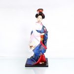 Boneca decorativa de estilo oriental / japonês(Ningyo), figurando uma Geisha tradicional. Representada em pé sobre base retangular de madeira escurecida, em pose clássica com as mãos recolhidas sob as longas mangas do traje. Apresenta cabeça moldada em gesso ou composição com acabamento acetinado límpido, exibindo feições finamente pintadas à mão com olhos amendoados e lábios carmim em formato de coração; penteado tradicional volumoso(nihongami) elaborado em fios sintéticos pretos, ricamente ornamentado por uma flor de tecido lilás na lateral e fitas vermelhas com fios dourados entrelaçados no topo do coque. Traja indumentária típica sobreposta composta por quimono  de mangas largas em tecido brocado azul-royal com padronagem floral e de folhagens fênix com fios metalizados furtacor. Destaca-se na manga direita caída uma expressiva estampa de garça branca de asas estendidas, sobrevoando ramagens floridas policromadas de folhas de plátano; traje complementado por subquimono em tons de rosa e branco, saia inferior vermelha e uma larga faixa abdominal(obi) rígida em tecido dourado com padronagem brocada de escamas ou ondas concêntricas em seu reverso. Medidas: Alt.: x Comp.: x Larg.: cm.