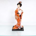 Boneca japonesa tradicional(Ningyo), figurando uma Geisha clássica. Representada em pé sobre base quadrada de madeira escurecida, em pose frontal. Apresenta cabeça moldada em gesso ou composição(gofun), com feições pintadas à mão ressaltando os olhos com pálpebras marcadas e lábios carmim bem delineados, penteado tradicional suntuoso(nihongami) elaborado em fios sintéticos pretos, ornamentado por presilha, fitas vermelhas com fios dourados e flores artificiais na cor azul, traja indumentária típica sobreposta composta por quimono de mangas largas em tecido brocado de um vívido tom laranja, inteiramente decorado por tramas metalizadas douradas e prateadas figurando crisântemos e motivos florais da tradição japonesa, traje complementado por subquimono branco e vermelho, além de larga faixa abdominal(obi) rígida em tecido preto com brocados dourados imitando padronagem de ondas em seu reverso. Com ambas as mãos em cerâmica moldada,  segura à frente do corpo um tradicional leque de mão decorado por uma estampa cromolitográfica retratando outra geisha clássica portando um leque dobrável, acompanhada de caracteres caligráficos em japonês. Apresenta descolamento do cabelo no topo da cabeça, necessita reparo, vide foto. Medidas: Alt.: x Comp.: x Larg.: cm.