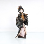 Boneca estilo japonesa tradicional(Ningyo), figurando uma Geisha clássica. Representada em pé sobre base quadrada de madeira escurecida. Apresenta cabeça moldada em gesso ou composição(gofun), com feições pintadas à mão detalhando olhos amendoados e pequenos lábios vermelhos, penteado tradicional elaborado em fios sintéticos pretos amarrados em coque alto, ornamentado por presilhas de flores artificiais brancas e roxas, e contas esféricas nas cores rosa e bege. Traja indumentária típica sobreposta composta por quimono de mangas largas em tecido texturizado escuro com efeito salpicado brilhante e metalizado em tons de prata, verde e azul, traje complementado por subquimono em tom rosa, gola bege e uma larga faixa abdominal(obi) rígida em tecido ocre-dourado texturizado, arrematada por um grande laço em seu reverso. Medidas: Alt.: x Comp.: x Larg.: cm.