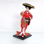 Boneca estilo japonesa tradicional(Ningyo), figurando uma Geisha clássica. Representada em pé sobre base quadrada de madeira pintada de preto, em pose de dança dinâmica com o quadril sutilmente projetado e as pernas envoltas pelo panejamento do traje. Apresenta cabeça moldada em gesso ou composição(gofun), com feições pintadas à mão ressaltando os olhos com delineado delicado e lábios carmim bem definidos, penteado tradicional elaborado em fios sintéticos pretos, guarnecido lateralmente por ornamentos circulares dourados em filigrana e fixado sob um grande e imponente chapéu plano de aba larga tradicional na cor preta, ricamente ornamentado no topo por debruns dourados e uma estampa radiada de folhagens espiraladas em tom creme com miolo florido saliente. Traja indumentária típica sobreposta composta por quimono de mangas largas em tecido brocado de um vívido tom vermelho-escarlate, inteiramente decorado por folhagens esmeralda e crisântemos tridimensionais tecidos com fios metalizados dourados e prateados, traje complementado por subquimono branco e rosa, e uma larga faixa abdominal(obi) rígida em tecido preto com brocados dourados em padronagem de ondas concêntricas em seu reverso. Com a mão direita erguida, a figura empunha um leque dobrável fechado inteiramente recoberto por película dourada texturizada com discretos grafismos vegetais incisos. Medidas: Alt.: x Comp.: x Larg.: cm. Apresenta colagem com fita adesiva no rosto, demanda pequeno reparo, vide foto.