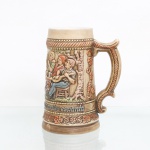 Caneca de chope estilo germânico em faiança moldada em alto-relevo e pintada à mão, produzida pela Ceramarte Ltda(Rio Negrinho-SC) c.1970-1985.Corpo em tom marrom-ocre acetinado com cena de camponeses a confraternizar numa taverna. Apresenta na base a inscrição em letras góticas: "Quem bebe e canta - seus males espanta". As bordas e a alça imitam cordas trançadas com relevos de lúpulo e videiras. No verso inferior, exibe a marca carimbada da Ceramarte e o número de molde 10 inciso. Medidas: Alt.: x Comp.: x Larg.: cm.