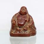 Escultura representando Buda Risonho(Hotei / Budai) executado em faiança moldada e esmaltada. Representado sentado em pose relaxada(asana) sobre base moldada que acompanha o contorno de suas vestes. É retratada em sua fisionomia característica com calvície proeminente, lobos auriculares alongados, feições risonhas e grande abdômen descoberto, simbolizando magnanimidade, satisfação e abundância; corpo recoberto por espesso esmalte translúcido de tom marrom, que escorre pelas rentrâncias criando sombreados escuros e destacando as dobras do panejamento de suas vestes, superfície apresenta um craquelê homogêneo sob o vidrado. Base de fundo vazado, interior oco. Medidas: Alt.: x Comp.: x Larg.: cm.