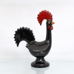 Escultura decorativa em faiança moldada e esmaltada figurando o tradicional Galo de Barcelos, representada em sua modelagem clássica estilizada de perfil, assentada sobre base circular alteada em pedestal de perfil anelar. Exibe corpo inteiramente recoberto por espesso esmalte monocromático na cor preta de alto brilho, crista arqueada e a barbela esmaltadas em vermelho, olhos  com pintura manual nas cores laranja e preto, e o bico cônico em tom ocre, cauda serrilhada características do icônico símbolo da identidade portuguesa. Fundo liso vidrado sem marcação de manufatura. Medidas: Alt.: 27,5 x Comp.: 20 x Larg.: 12,5cm