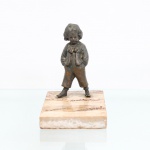 Escultura representando Menino em bronze sobre base de mármore. Escultura decorativa figurando um menino com as mãos nos bolsos, executada em bronze fundido com pátina escura. Representada em pé, com traje de época composto por colete ornamentado, camisa de mangas bufantes com laço estruturado no pescoço, calças curtas e botas de amarrar. Toda a composição assenta sobre uma robusta base retangular em mármore. Medidas: Alt.: x Comp.: x Larg.: cm.