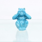 Figura de macaco Não vejo o mal, em cerâmica esmaltada. Representação de macaco sentado, com ambas as mãos cobrindo os olhos, correspondente a Mizaru, um dos Três Macacos Sábios da tradição iconográfica japonesa. Modelagem moldada, revestida por esmalte azul-turquesa, com apontamentos esverdeados nas extremidades. A composição integra a máxima associada a Mizaru, Kikazaru e Iwazaru - respectivamente não ver o mal, não ouvir o mal e não falar o mal. Medidas: Alt.: x Comp.: x Larg.: cm.