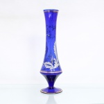 Vaso solitário em vidro azul cobalto, corpo alongado de seção circular, perfil cônico com colo estreitado e boca discretamente aberta, elevado sobre haste curta e pé circular. Ornamentação de caráter vegetalista aplicada em esmalte, composta por folhagens estilizadas e volutas, complementada por filetes dourados na boca, no terço inferior e na base. Medidas: Alt.: x Diam.: cm.