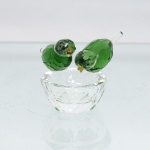 Porta-aliança em cristal facetado, composição formada por casal de pássaros pousados sobre recipiente circular representando ninho. Aves executadas em cristal verde, com cabeças incolores facetadas, olhos aplicados e bicos em tonalidade âmbar; caudas alongadas em vidro transparente. Base em forma de pequena taça, com paredes e bordo facetados. Sem marca de manufatura. Medidas: Alt.: x Diam.: cm.