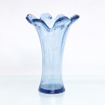 Vaso executado em vidro soprado, corpo tubular, tonalidade azul-claro translúcido com inclusões de bolhas decorrentes do processo de fabricação. Boca expandida e conformada manualmente em seis lóbulos, formando bordo ondulado; base circular espessada em tonalidade azul de maior saturação. Medidas: Alt.: x Diam.: cm.