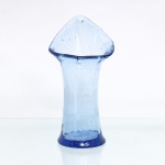 Vaso em vidro soprado, tonalidade azul-claro translúcido, de corpo tubular afunilado na região mediana e base circular espessada. Terminação superior aberta e modelada a quente, configurando boca assimétrica de contorno triangular. Presença de inclusões de bolhas e irregularidades próprias do processo de sopro manual. Medidas: Alt.: x Diam.: cm.