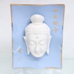 Placa decorativa em porcelana esmaltada, de formato retangular vertical e perfil levemente ondulado, com fundo em azul-claro e contorno dourado. Ao centro, aplicação em alto-relevo representando a cabeça de Guanyin (Kuan Yin) em porcelana branca, com olhos semicerrados, lóbulos auriculares alongados, cabelos reunidos em coque e ornamento frontal com motivo floral. Inscrições em caracteres chineses dourados, incluindo Baiyi Guanyin, denominação de Guanyin de Vestes Brancas, manifestação da bodhisattva da compaixão na tradição budista chinesa. Verso com cordão para suspensão. Medidas: Alt.: 24,5 x Comp.: 19 x Larg.: 7,5cm