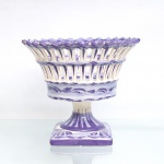 Fruteira de pé em faiança, corpo composto por taça circular vazada, estruturada em duas ordens de reservas verticais, bordo recortado em sequência angular e haste central sobre base quadrangular. Decoração pintada à mão em monocromia violeta, com frisos vegetalistas, filetes e composição floral no fundo da cuba. Base com inscrição manuscrita sob o vidrado . Apresenta perdas pontuais de esmalte no bordo. Medidas: Alt.: 17,5 x Diam.: 20cm.
