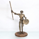 Escultura figurativa executado em bronze fundido com pátina dourada, representando a personagem literária Dom Quixote de la Mancha, meados do século XX. Apresenta-se envergando armadura composta por couraça, grevas e gorjal, além da bacia de barbeiro utilizada como elmo(Elmo de Mambrino). O braço destro eleva uma lança longa, enquanto a mão esquerda empunha um escudo circular côncavo portando uma espada embainhada presa à guarnição posterior. Assente sobre plinto circular integrado de contorno irregular. Apresenta fratura  na junção do pescoço. Medidas: