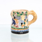 Caneca decorativa em faiança moldada e policromada, de procedência japonesa, período Showa, circa 1930-1950. Apresenta corpo de secção cilíndrica com decoração em alto-relevo de caráter pictórico e folclórico, ilustrando casais em trajes tradicionais europeus (estilização de festividade camponesa ou taverna) inseridos em cenário arquitetônico e vegetal com ramagens e frutos suspensos. Alça lateral mimetiza uma estrutura orgânica de tronco ou galho retorcido com acabamento vidrado ocre; base circundada por friso de arcos concêntricos em relevo sobre fundo azul, assentando sobre rodapé esmaltado de tonalidade preta. Verso da base ostenta a marca de exportação carimbada sob o vidrado: "Made in Japan". Apresenta craquelado característico do material. Medidas: Alt.: x Comp.: x Larg.: